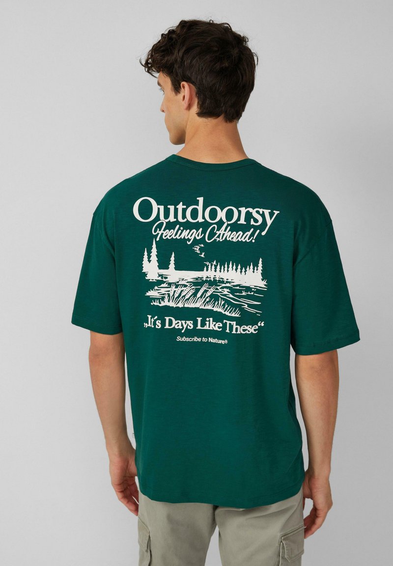 Groene katoenen t-shirt met korte mouwen, waarop witte tekst staat en een natuurthema grafiek op de achterkant, met bomen en water.