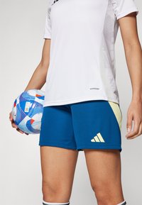 Mėlynos sportinės šortai su geltonais akcentais ir Adidas logotipu, derinami su balta sportine marškinėliais, kuriuose yra tekstūruota šoninė dalis. Laikoma raštuota futbolo kamuolys.
