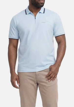 Homme portant un polo à manches courtes bleu clair avec un bord marine sur le col et les manches, associé à un pantalon beige.