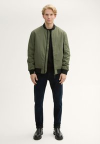 Olivgrön bomberjacka med svarta muddarna, bärs över en svart skjorta; kombineras med mörk denimjeans och svarta ankelboots.