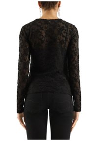 Schwarzes Langarmoberteil mit floralen Spitzenverzierungen, durchsichtigem Stoff und körperbetontem Design. Kombiniert mit dunklen Jeanshosen.