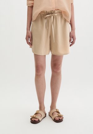 Person trägt beigefarbene Shorts mit Kordelzug, ein pfirsichfarbenes Hemd und beigefarbene Sandalen mit Schnallen und steht vor weißem Hintergrund.