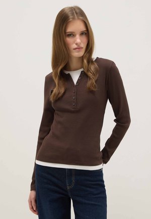 Jeune femme aux longs cheveux bruns portant un haut henley boutonné à manches longues marron foncé sur un chemisier blanc et un jean bleu foncé.