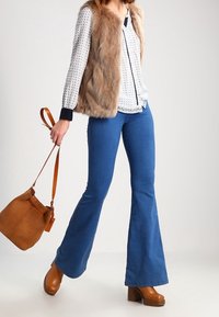 Gilet in pelliccia sintetica marrone sopra una camicia bianca con motivo, abbinato a jeans blu a zampa d'elefante e scarpe con tacco platform marroni. Include una borsa marrone.