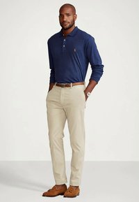 Sötétkék hosszú ujjú galléros póló kis logóval, bézs chinos nadrág, barna öv és barna mokaszin. A személy zsebében tartja a kezét, áll.