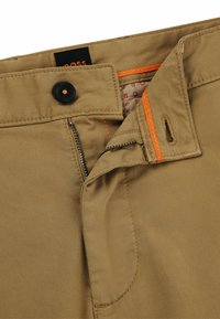 Beige trousers with a zip and button closure, featuring contrasting orange stitching and an inside patterned lining. Smooth fabric texture.