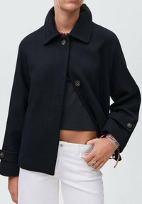Femme portant une veste noire à boutons sur un top court noir et un pantalon blanc, avec des bracelets et des bagues colorés sur sa main.