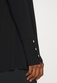 Czarnoskóra ręka spoczywa na boku, ubrana w długą czarną koszulę z trzema białymi guzikami na mankiecie, na jednolitym tle.