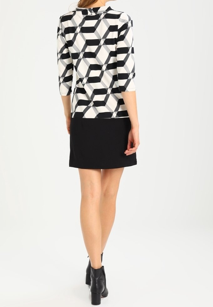 Trekvartärmad topp i ett svart och vitt geometriskt mönster, i kombination med en svart kjol. Tyget har en slät textur och visar en modern design.