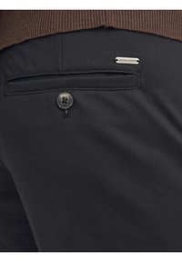 Zwarte broek met een achterzak voorzien van een zwarte knoop en een metalen logo-accent. De stof lijkt glad te zijn met een lichte glans.