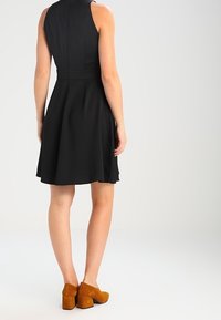 Black sleeveless dress with a fitted bodice and flared skirt. Worn with brown suede block-heel shoes. Smooth texture.