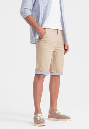 Khaki shorts dengan cuff yang digulung, dilengkapi dengan saku samping dan ritsleting. Dipadukan dengan kemeja lengan panjang bergaris biru muda dan sepatu kets beige.