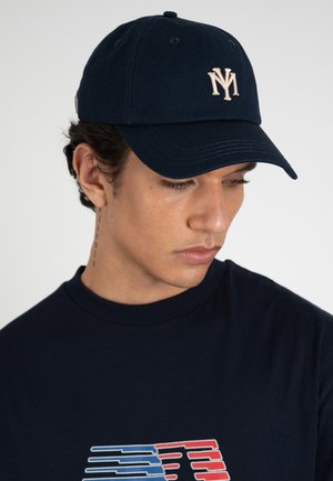 Jonge man met een marineblauwe pet met beige NY-logo en een marineblauw shirt met rode en blauwe afbeelding, kijkt omlaag tegen een effen achtergrond.