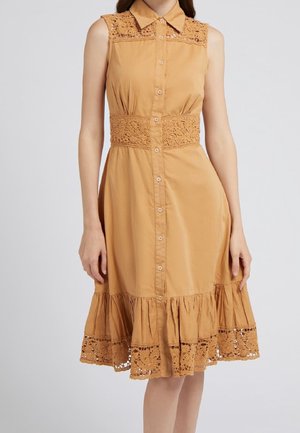 Robe beige sans manches avec fermeture boutonnée devant, détails en dentelle sur le col, la taille et l'ourlet, portée par une personne debout sur un fond uni.