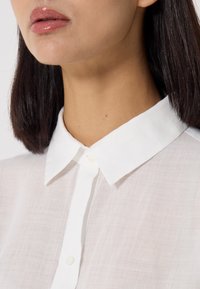 Camicia bianca con bottoni realizzata in tessuto trasparente, con colletto classico. Presenta un design pulito e una texture sottile, con bottoni visibili.