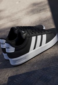 Baskets noires Adidas avec des rayures blanches et des semelles blanches posées sur une surface grise avec des ombres légères.