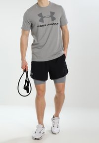 Camiseta gris de manga corta con un logo negro de "Under Armour", combinada con pantalones cortos negros sobre mallas de compresión grises, sosteniendo una banda de resistencia.