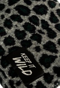 Textil gris con un patrón de leopardo en negro y verde oscuro, y una textura suave y peluda. Incluye una etiqueta negra con el texto blanco "MANTENLO SALVAJE."