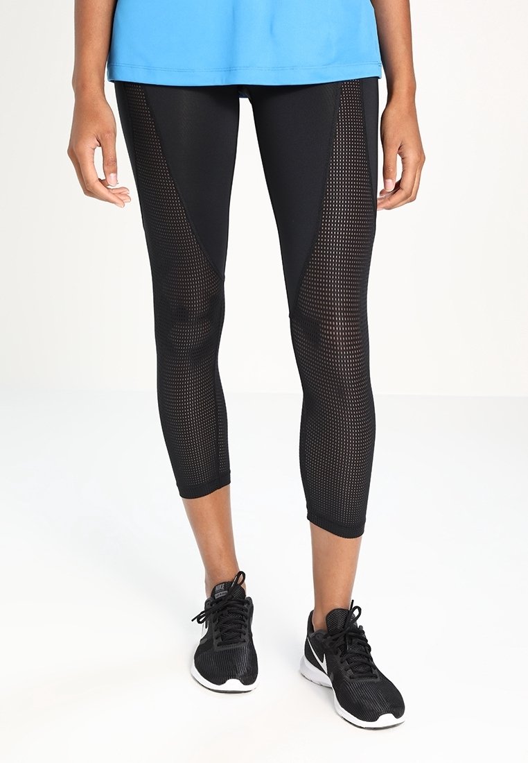 Czarne legginsy sportowe z siateczkowymi panelami, o skróconej długości i gładkiej fakturze, w zestawieniu z czarnymi butami sportowymi. Niebieska bluzka widoczna na górze.
