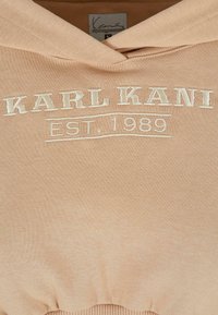 Felpa beige con logo ricamato "KARL KANI" e "EST. 1989" sottostante. Presenta un cappuccio con cordino e orlo a coste. Tessuto di cotone morbido.