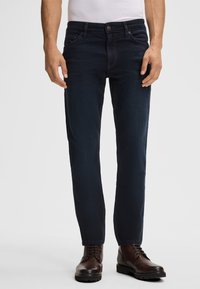 Jeans slim-fit blu scuro con un design classico a cinque tasche, realizzati in un materiale denim liscio e leggermente elastico. Stivali marroni con lacci completano il look.