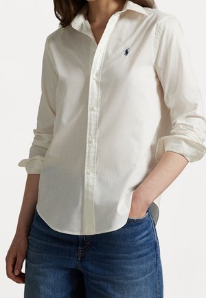 Mujer con una camisa blanca abotonada con las mangas remangadas y vaqueros azules, una mano en el bolsillo, mostrando un pequeño logo bordado en el pecho.