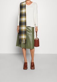 Pull à col en V blanc, jupe en cuir verte, écharpe à carreaux jaune et noire, chaussures à talons carrés marron, et sac seau marron.