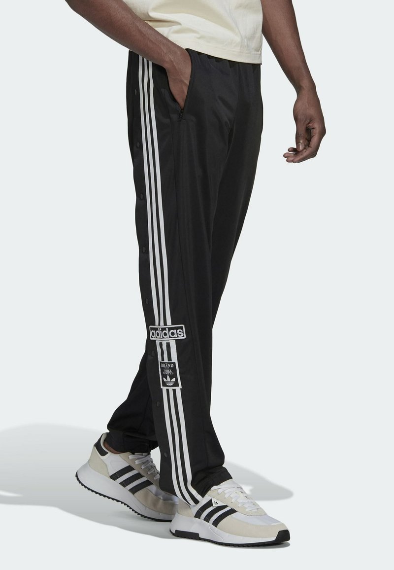 Man wearing black Adidas track pants with white stripes and logos, paired with white Adidas sneakers with black stripes.