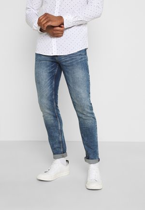Homme debout portant un jean bleu retroussé, des baskets blanches et une chemise blanche boutonnée avec de petits motifs noirs.