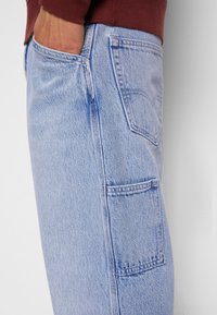 Main dans la poche d'un jean denim bleu clair avec poches latérales et arrière, porté avec un haut à manches longues rouge foncé visible à la taille.