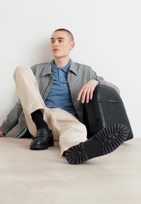 Sac à dos noir en tissu texturé, veste à carreaux grise, chemise polo bleue, pantalon beige clair et chaussures noires sans lacets sur un sol neutre.