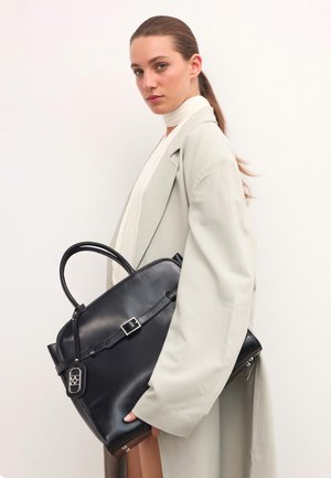 Sac à main en cuir noir avec une forme structurée, doté d'une poignée supérieure, de détails à boucle et d'une étiquette amovible, tenu contre un manteau de couleur claire.