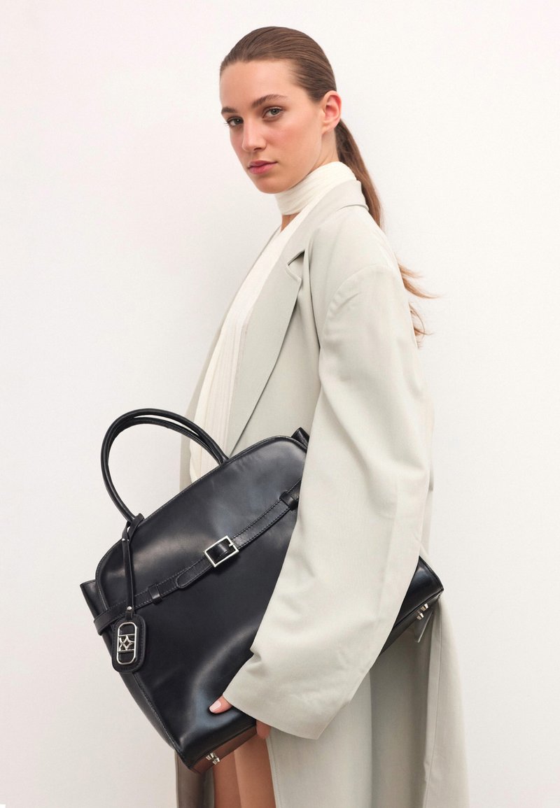 Melna ādas rokassoma ar strukturētu formu, kurai ir augšējā roktura, sprādze un noņemama birka, turēta pret gaismas krāsas mēteļa.