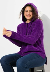 Felpa viola con cappuccio in tessuto texturizzato, polsini a costine e tasca frontale. Indossata sopra jeans blu, modello seduto su uno sgabello nero.