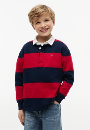 Garçon blond souriant, main dans la poche, portant un maillot de rugby rayé rouge et marine et un jean bleu clair sur fond blanc uni.
