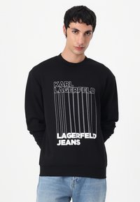 Svart hettegenser laget av mykt stoff som har fet hvit grafisk tekst "KARL LAGERFELD JEANS" og vertikale linjer på forsiden.