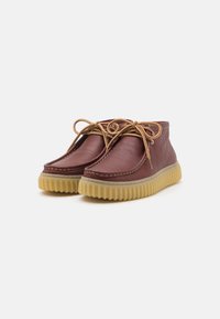 Brown leather ankle boots with beige rubber sole and textured finish. Features rounded toe and contrasting laces. Stitched detailing visible.