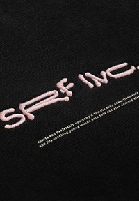 Czarna tkanina z różowym haftowanym napisem "SRF INC." nad mniejszym białym tekstem. Struktura wydaje się gładka z lekko prążkowym wykończeniem.