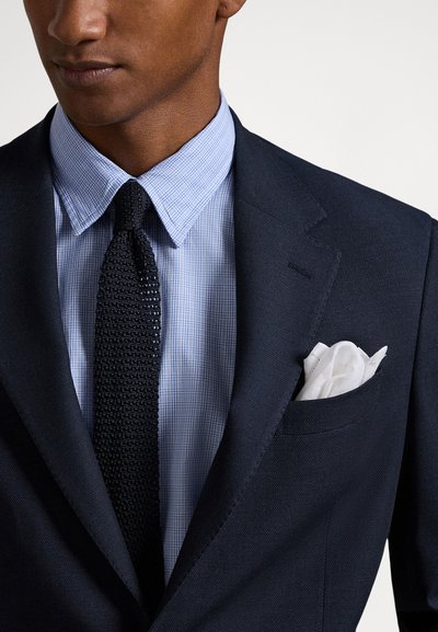 Vīrietis, kurš valkā tumši zilu žaketi, gaiši zilu rūtainu kreklu, melnu adītu kaklasaiti un baltu kabatas lakatiņu žaketes kabatā.