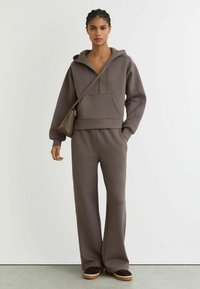 Sudadera marrón con capucha, bolsillo delantero y cremallera, combinada con pantalones de pierna ancha a juego. Presenta una textura suave y un ajuste relajado.