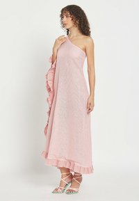 Mujer con cabello rizado que lleva un vestido rosa de un solo hombro hasta el suelo con bordes con volantes y sandalias de tacón alto con tiras.