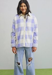 Fleece button-up jack met een paars en crèmekleurig geruit patroon. Heeft een puntige kraag en zijzakken. Gepaard met versleten lichte jeans.