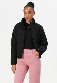 Svart pufferjacka med hög krage, dragkedjestängning och vadderad yta, i kombination med ljusrosa jeans med knappstängning.