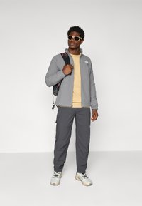 Pilka zip-up striukė, smėlinis marškinėliai, pilki krovinių kelnės ir baltos sportinės bateliai. Modelis laiko juodą kuprinę ir vilki baltus akinius nuo saulės.