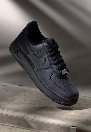 Sort læder Nike Air Force 1 sneaker med perforeret tå, sorte snørebånd og metal mærkat med mærke på snørebåndene, vist på betonoverflade.