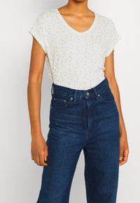 Camiseta blanca de manga corta y escote en V con lunares dorados, combinada con jeans de talle alto en un azul oscuro. Material de algodón con corte relajado.