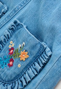 Világoskék farmerdzseki, amelynek fodros zsebe színes virágos hímzéssel díszített pink, sárga, kék és zöld fonalakkal.