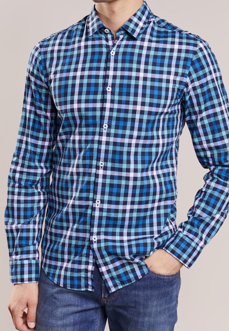 Homme portant une chemise à carreaux bleue, noire et blanche à manches longues et un jean bleu, une main dans la poche.