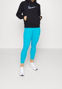 Osoba v černé mikině Nike s vzorovaným logem, jasně modrých legínách Nike a bílých sportovních botách, stojící na jednobarevném pozadí.