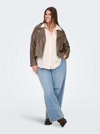 Lächelle Frau mit langen braunen Haaren, die eine braune Lederjacke, eine cremefarbene Bluse, hellblaue Jeans und dunkelbraune Stiefel trägt und mit einer Hand in der Hosentasche steht.